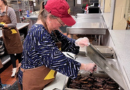Julee Gard, wearing a red cap, a navy blue and white striped shirt, and a brown apron, is focused on serving sausages at a soup kitchen in Joliet, Illinois, alongside her University of St. Francis colleagues.