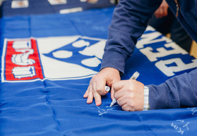 A close-up of a senior staff member signing his name with a white marker on a blue Penn flag. It will be flown above the construction project over the next few months.