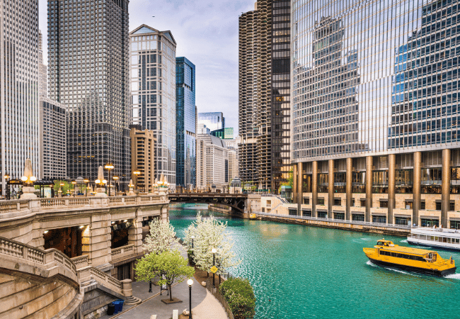 An iconic view of Chicago, Illinois, with the river (center) curving through downtown. It’s a beautiful sunny afternoon. A bright yellow sightseeing cruise boat enters the frame from the lower right-hand corner, and skyscrapers tower over on both sides. In the distance, we see several iconic bridges crisscrossing over the shimmering, bright green water.