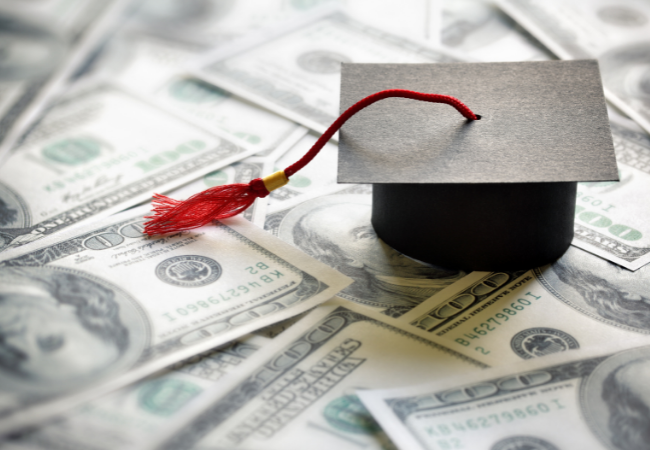 A photo illustration of a mini mortarboard (graduation cap) sitting on top a stack of $100 bills