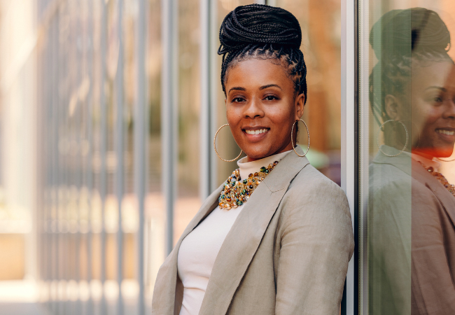 Assistant Professor Ericka Weathers pictured outside Penn GSE on a sunny spring afternoon. She is leaning against a reflective glass wall as she smiles at the camera.