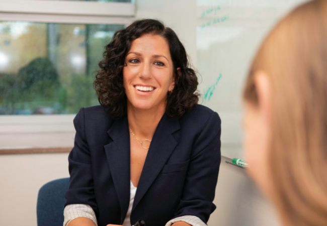 Dr. Katharine O. Struck casually seated at a conference table, facing the camera, with a whiteboard and windows onto a leafy campus behind her. She is smiling and listening to a student who is facing her, and who has their back to the camera.