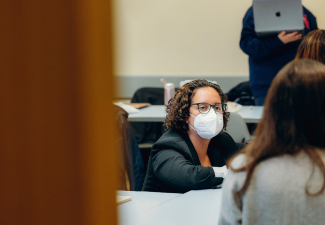 A photo in a classroom. Professor Sade Bonilla is leaning down at desk level to talk with two of her seated students whose backs are to the camera. They are all deep in conversation.