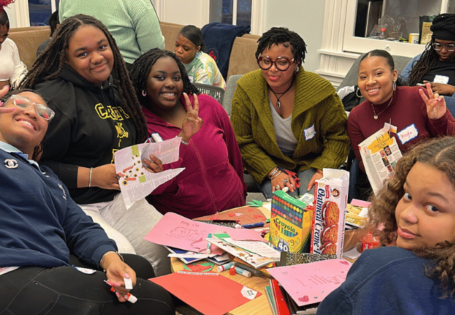 A group of teenage high school students hanging out during a recent Black Girls Literacies Project meeting. Everyone is casually dressed and comfortable. They’re seated around a coffee table piled high with school boxes, art supplies and snacks. They’re in the middle of doing a project. Everyone is happy and smiling, and several of the students are making the peace sign with their hands. It looks fun and friendly.