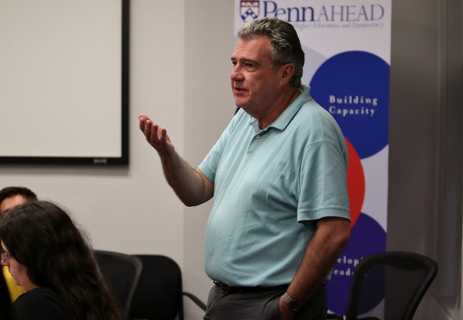 Alan Ruby teaching a class. He’s standing in a classroom, talking, while facing tables of students listening and taking notes. A banner behind him promotes the Penn AHEAD program, where Ruby is a senior fellow.