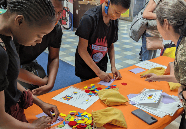 Students, teachers and facilitators are pictured in a school during a recent math festival organized by Penn GSE’s Responsive Math Teaching project. They’re gathered around a table with games and activities spread out. Three middle school-age students in the foreground are completing an activity while an instructor looks on, offering advice.