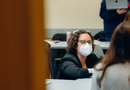 A photo in a classroom. Professor Sade Bonilla is leaning down at desk level to talk with two of her seated students whose backs are to the camera. They are all deep in conversation.