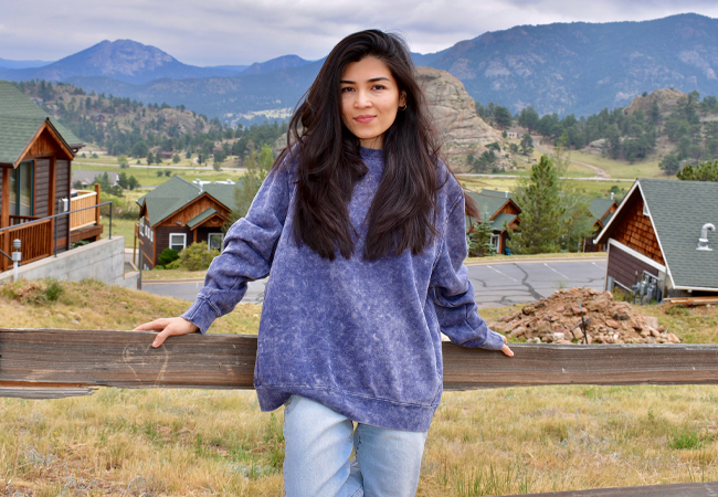 Yasmeen wears a purple sweatshirt and jeans as she leans on a fence with the RockyMountains behind her.