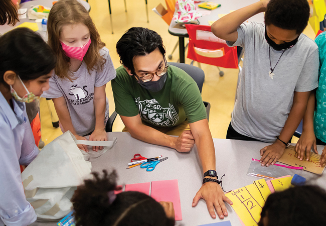 A seated teacher, wearing a face mask and glasses, is surrounded by several masked elementary students and reaches across a table to engage with a student's paper.