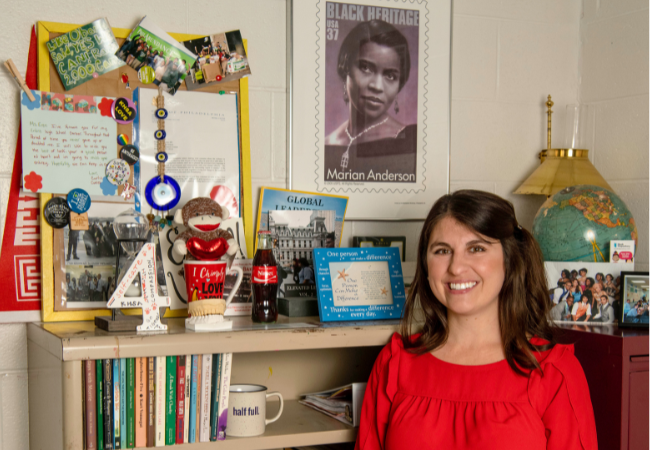 Nimet Eren, dressed in red attire, stands in her office surrounded by a collection of artifacts that reflect her journey as an educator.
