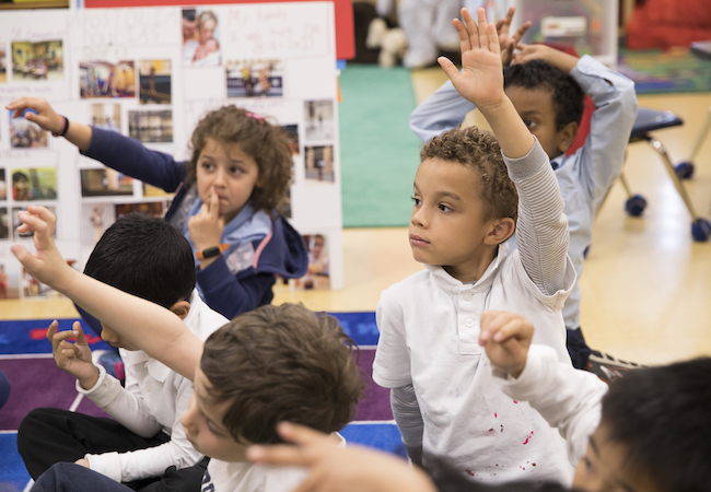 Children raise their hands while sitting on a mat in Kindergarten classroom