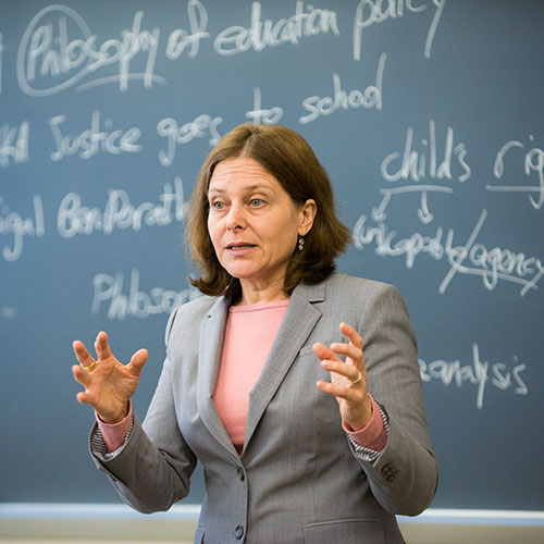 A professor addresses a class, standing in front of a blackboard with white chalk writings related to philosophy and education.