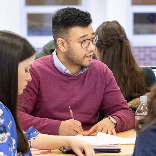 Two graduate students are sitting at a desk in a classroom, with other students seen in the background. One student takes notes while conversing, the other student looks down.