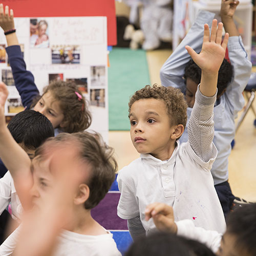 Elementary school children sit on the floor with their hands raised inside a colorful classroom.