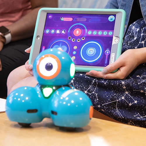 A blue robotic device with three spheres sits on a table. Behind it, an individual holds up a tablet device showing illuminated circles.