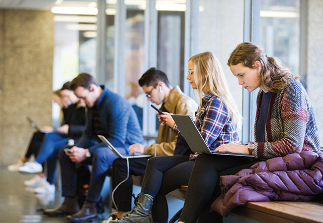 Several students are sitting next to each other in a well-lit space, working on their laptops and phones.