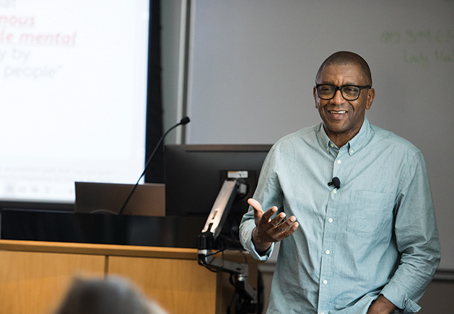 A smiling professor has his right hand raised midway and left hand in his pocket. He is wearing a blue shirt that has a lapel mic on it.