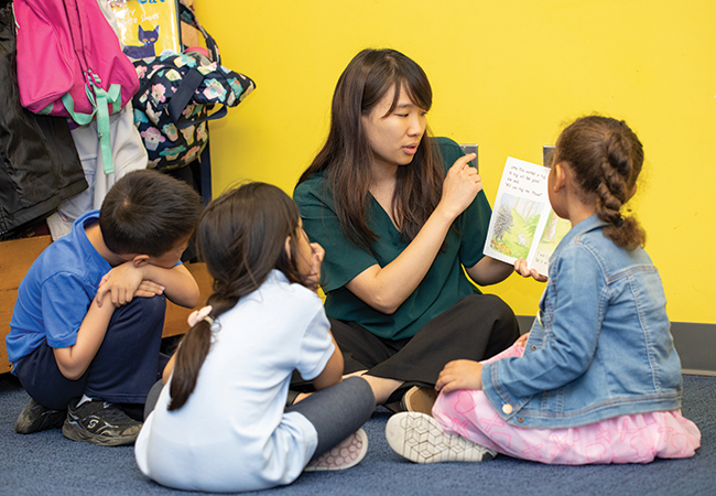 Three children are seated around a young woman in a classroom with yellow-colored walls and blue-colored carpet. They listen as the young woman narrates a story from a book she is holding.