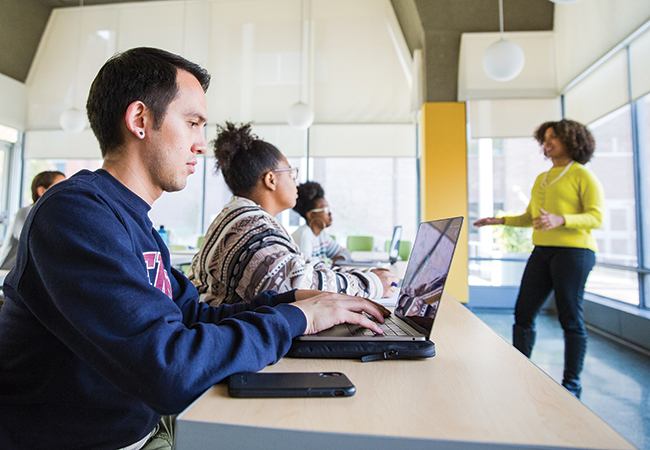 Three students are sitting inside a bright classroom which has a tall ceiling and pendant lights. They are working on their laptops and listening to the lecturer.