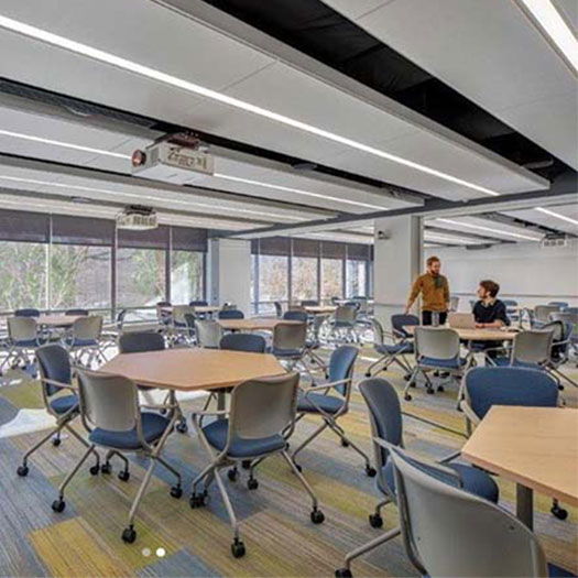 A large, well-lit classroom with long windows and high ceilings has many hexagon-shaped tables and revolving chairs. Two students are seen working and speaking with each other at a table in the far right