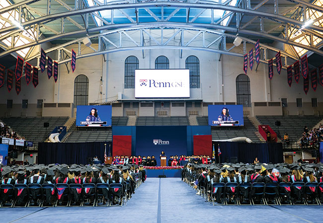 Inside Penn’s Palestra, graduates in caps and gowns are seated in rows facing the stage, where faculty are similarly dressed and seated and a speaker stands at a podium. Above the podium, a large screen reads “Penn GSE.”