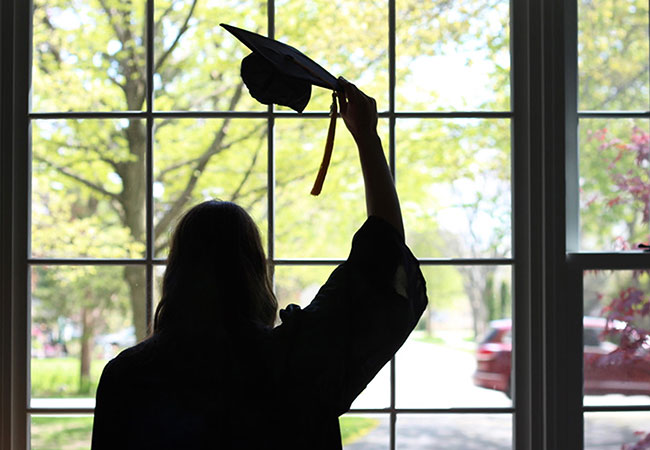 A person in a graduation gown appears in silhouette before a window, holding up a mortarboard with one hand