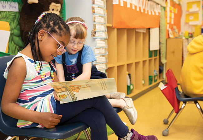 Two young girls in a colorful classroom sit on chairs reading from the same book