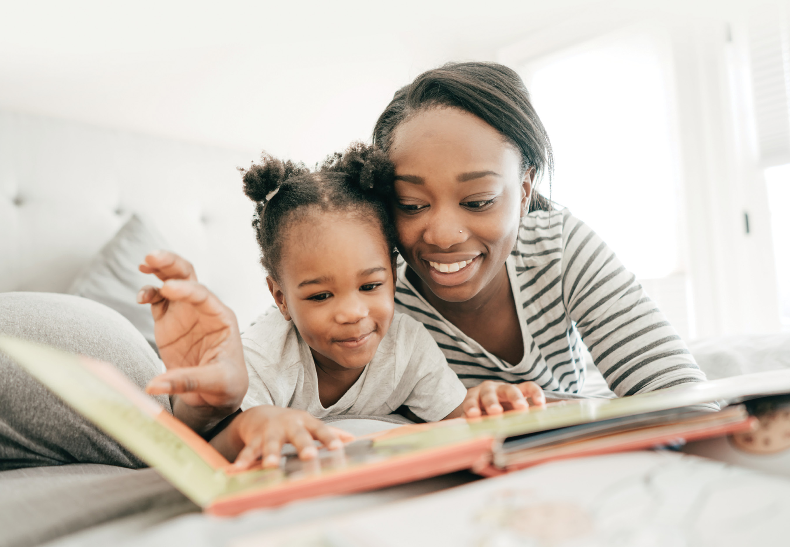 A young woman and a preschool girl with smiles on their faces are looking at a large hardcover book together.