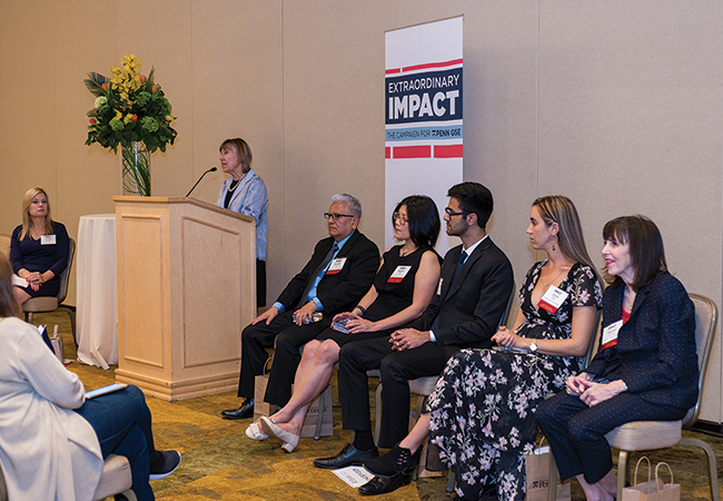 Dean Pam Grossman speaks at a podium in the front of the room. On either side of her are six men and women sitting panel-style in chairs, wearing professional attire with nametags.