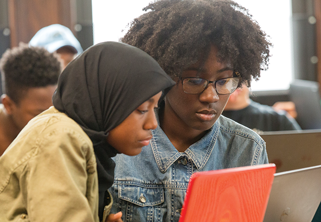 Two adolescent students in a classroom look down at a laptop screen