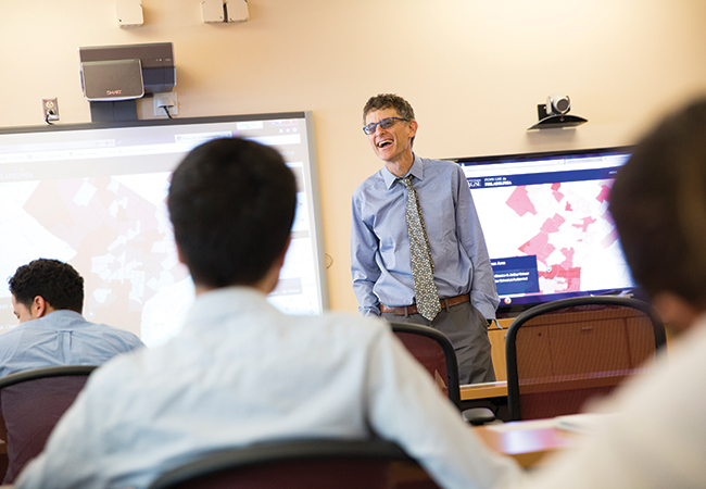 A professor wearing a blue shirt and patterned tie, smiles and addresses students in a classroom. Behind him are screens containing digital media.