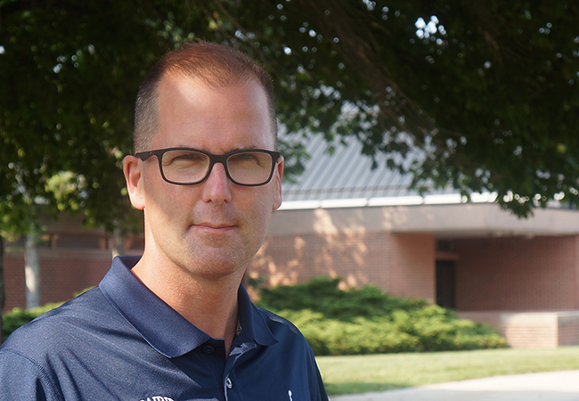 Nathaniel Coffman smiles in front of a school building wearing glasses and a basketball coach shirt.