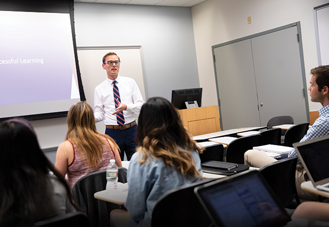 A man in professional attire is speaking in front of a classroom. Behind him is a projector displaying a PowerPoint presentation, and in front of him are several students.