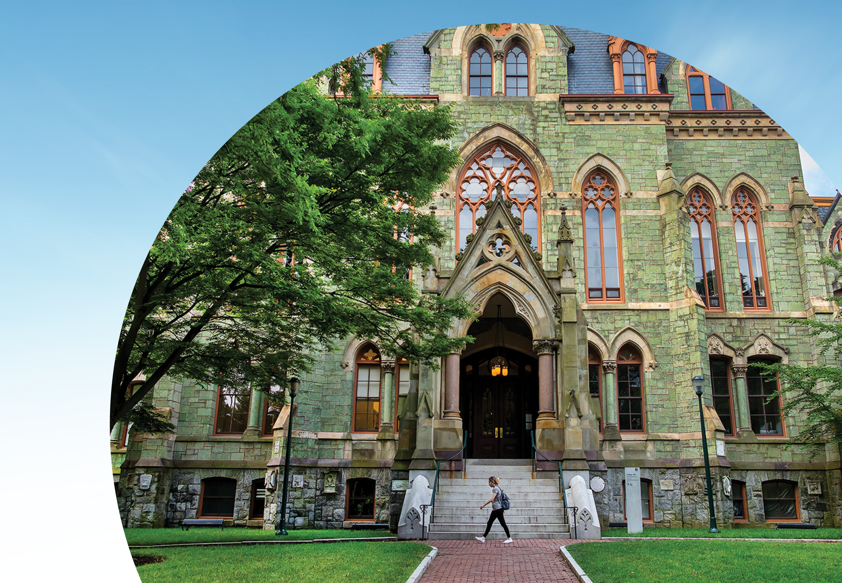 A student walks in front of College Hall building on the Penn campus., towards a leafy green tree. The photo is cropped with a semicircular edge against a blue-sky background.