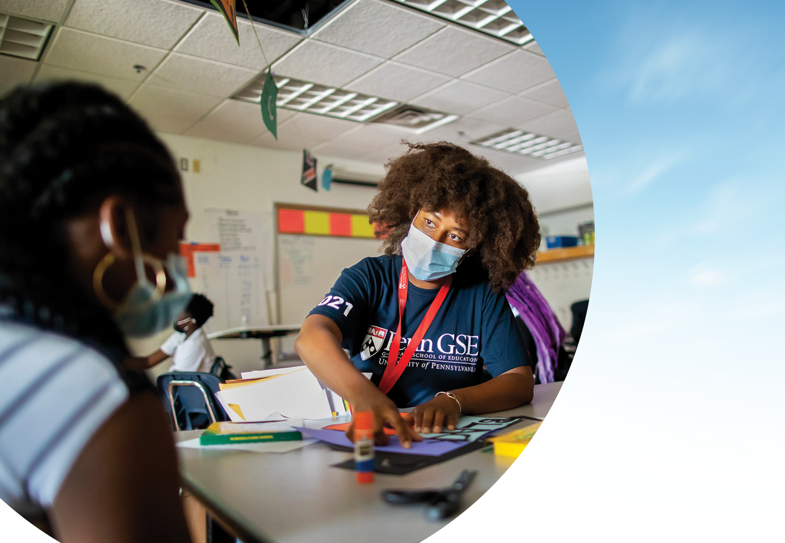 An instructor wearing a mask and a Penn GSE T-shirt is and engaged in conversation with a school student as they work on a project on a table. The photo is cropped with a semicircular edge against a blue-sky background.