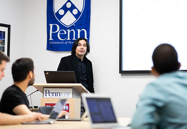 A professor wearing a suit speaks to students in a classroom. A blue Penn banner is behind him, and a Penn GSE logo is on the podium where he stands.