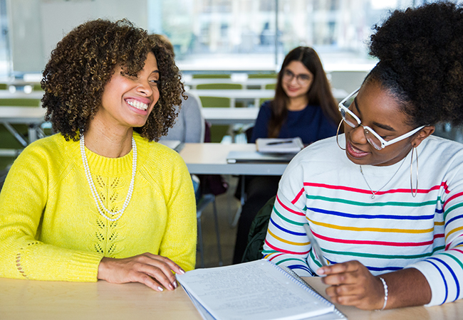 A professor and student wearing bright colors sit at a desk in a classroom with smiles on their faces, discussing academic material.