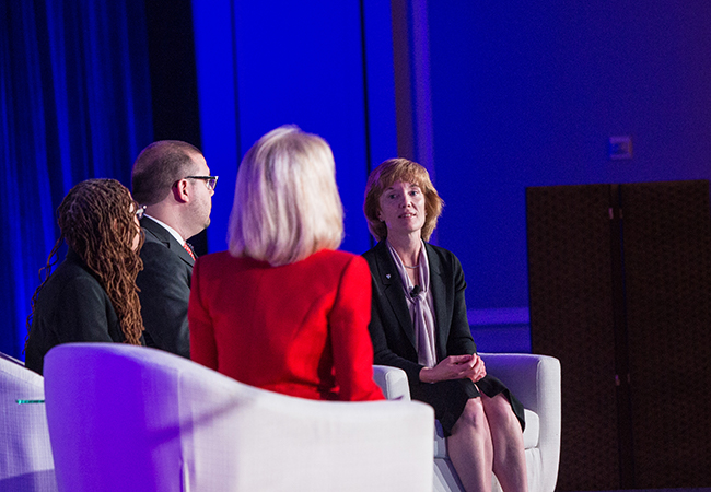 Four individuals in professional attire are seated in front of a blue curtain on a stage and engaged in conversation.