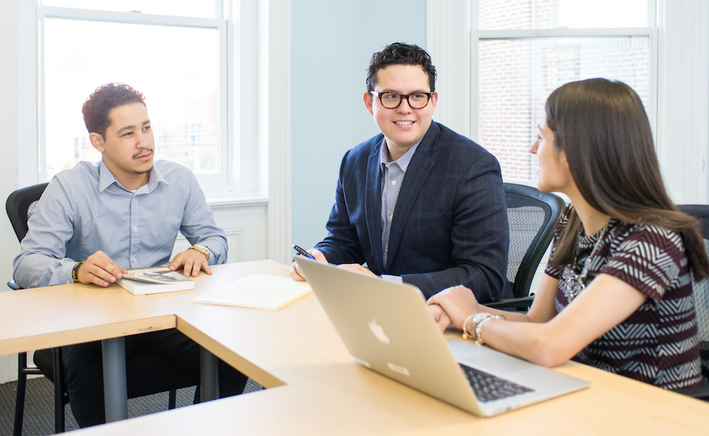 Three graduate students talking at a table with a laptop and book