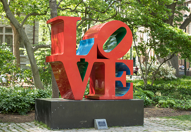 The iconic red LOVE sculpture appears amidst green foliage on Penn’s campus