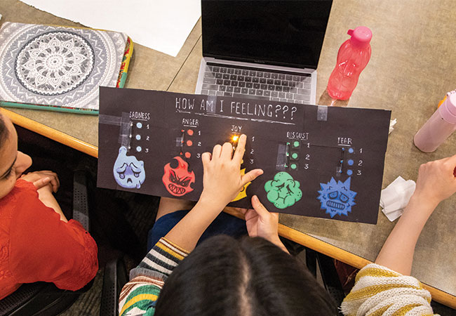 Aerial view of a student holding a colorful chart with a list of emotions and a light shining in the middle underneath the word “Joy.”