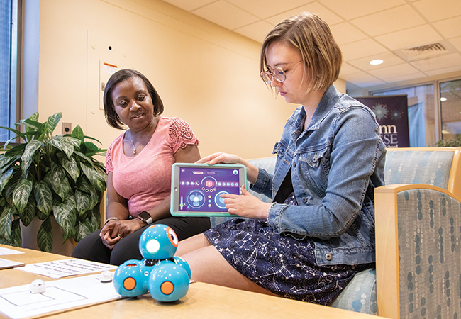 Two women sitting side by side demonstrate how to use a tablet device to operate a blue Dash Robot on the table in front of them