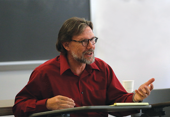 Photo of Michael Nakkula in a red button-up shirt and glasses sitting at a desk and teaching