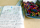 A folded composition book with children’s writing and drawings lies next to a tray of crayons 