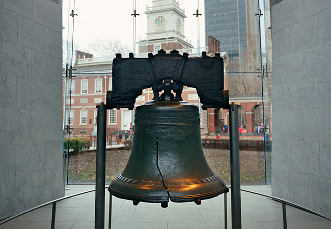 The Liberty Bell in Philadelphia appears between two gray walls, with traditional and modern buildings seen through a window behind it.