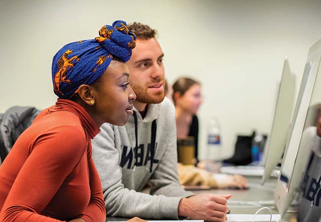 Two adult students sit side by side, smiling and looking at a screen in a computer lab.