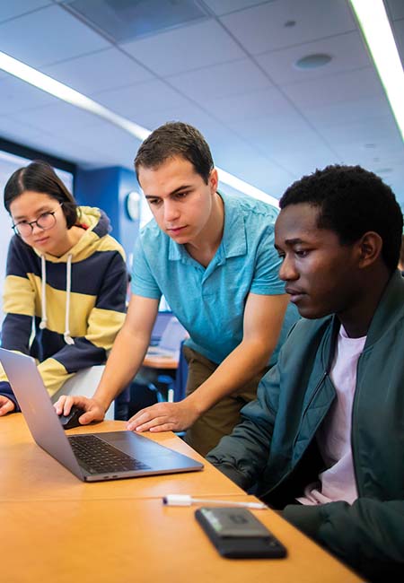 Three students look at an opened laptop screen inside a well-lit classroom.