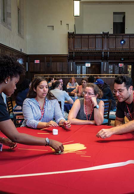 Adult students collaborate on a group task around a table, placing sticks on its surface.