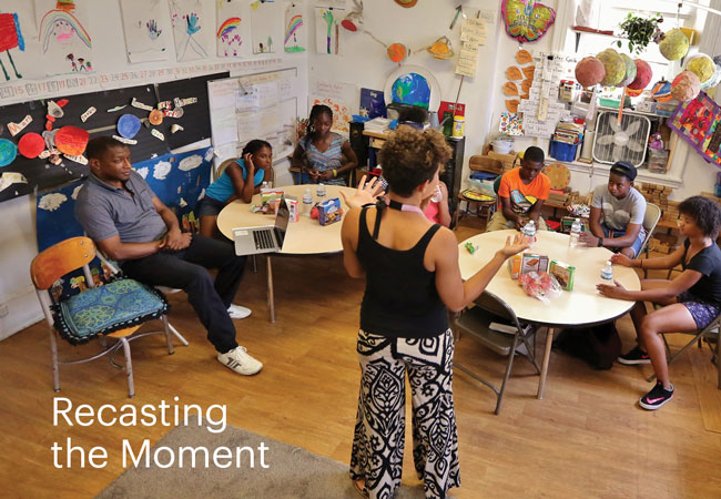 Elementary school students sit at round tables in a classroom. Behind them are colorful decorations, classroom projects, and school supplies. A woman stands at the front of the room and a man sits to the left. A headline on the image reads, “Recasting the Moment.”