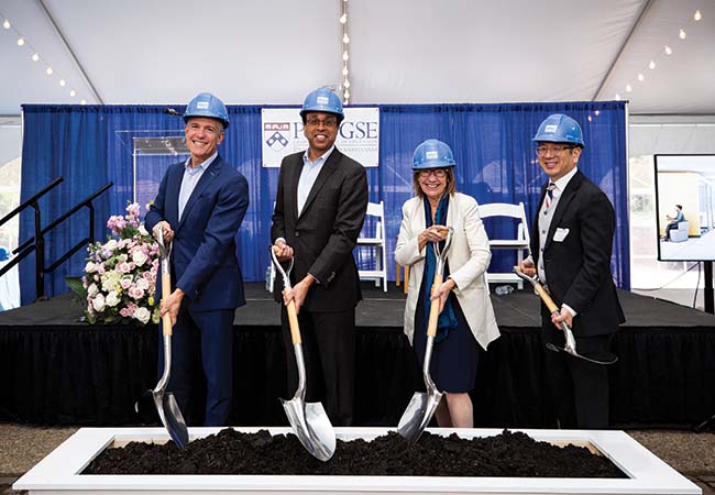 Four individuals standing next to each other and facing frontwards are wearing blue-colored hard hats and putting shovels into a pile of dirt.